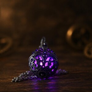 May include: A spherical metal pendant featuring an intricate, dark-toned filigree cage design. The interior of the pendant emits a vibrant, glowing purple light that shines through the decorative cutouts. A matching metal chain is attached to the top.