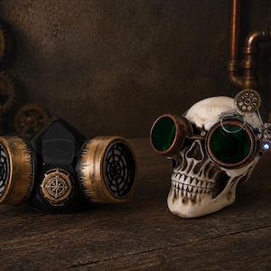 May include: A black respirator mask with metallic gold-toned filter covers and a central compass emblem, positioned next to a human skull replica wearing green-tinted goggles with a small gear detail and a single blue LED light attachment.