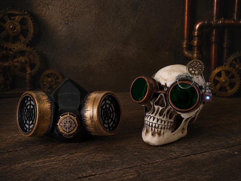 May include: A black respirator mask with metallic gold-toned filter covers and a central compass emblem, positioned next to a human skull replica wearing green-tinted goggles with a small gear detail and a single blue LED light attachment.