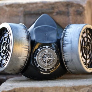 May include: A black gas mask with two silver filters and a compass design on the front. The mask is sitting on a stack of brown bricks.