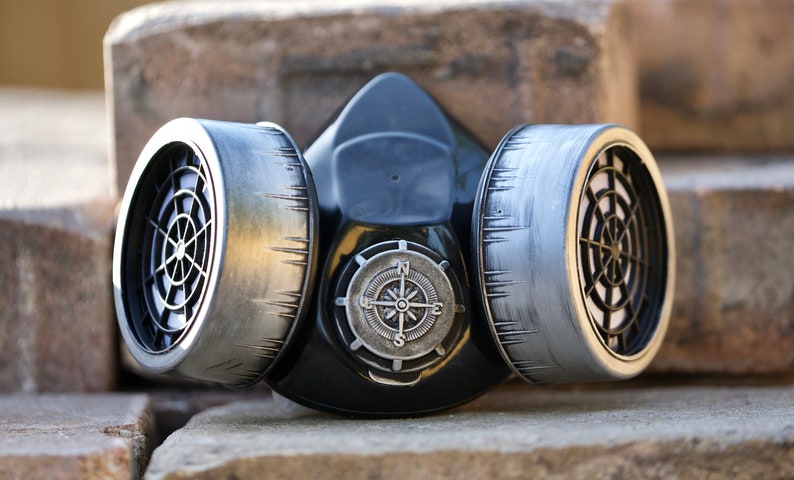 May include: A black gas mask with two silver filters and a compass design on the front. The mask is sitting on a stack of brown bricks.