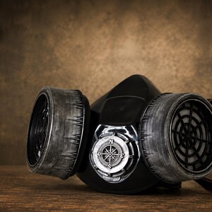 May include: A black respirator mask featuring two large, weathered grey cylindrical filters on the sides and a central circular silver compass emblem with cardinal directions N, S, E, and W clearly visible on the front face of the mask.