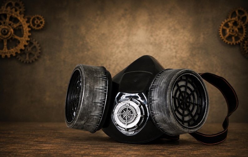 May include: A black respirator mask featuring two large, weathered grey cylindrical filters on the sides and a central circular silver compass emblem with cardinal directions N, S, E, and W clearly visible on the front face of the mask.