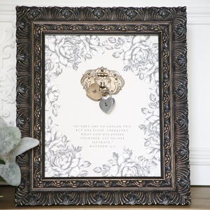 May include: A framed print with a floral design and a decorative lock with two heart-shaped charms. The frame is dark brown with ornate detailing. The print features a quote from Matthew 19:6. The background has a white and gray floral pattern.