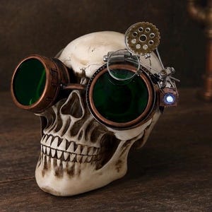 May include: A human skull replica wearing copper-toned goggles with dark green lenses. The goggles feature an attached mechanical gear mechanism, a small magnifying lens, and a tiny glowing blue LED light on the side of the frame.