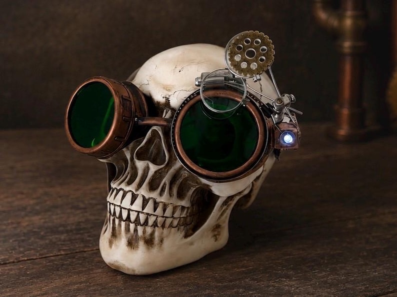 May include: A human skull replica wearing copper-toned goggles with dark green lenses. The goggles feature an attached mechanical gear mechanism, a small magnifying lens, and a tiny glowing blue LED light on the side of the frame.