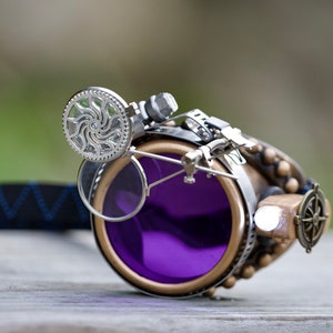 May include: Steampunk goggles with a purple lens, a silver gear detail, and a compass charm. The goggles are attached to a black strap.