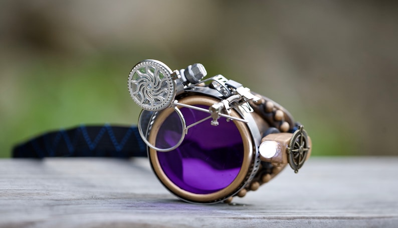 May include: Steampunk goggles with a purple lens, a silver gear detail, and a compass charm. The goggles are attached to a black strap.