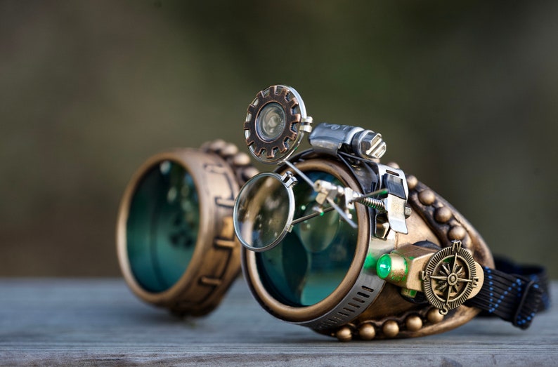 May include: Steampunk goggles with brass and copper accents, featuring a magnifying glass, a compass, and a green LED light. The goggles have a black elastic strap.