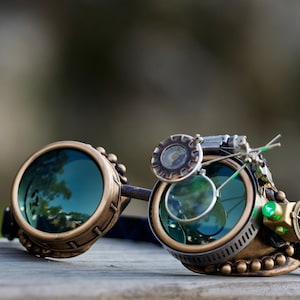 May include: Steampunk-style goggles with brass and silver accents. The goggles have round lenses with a green tint and a small magnifying glass attached to the side. There is a green light and a compass-like detail on the right side.