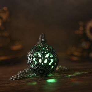 May include: A spherical pendant necklace featuring an intricate, dark metallic filigree cage design. The hollow interior emits a soft, glowing light in a pale green hue, visible through the ornate cutouts of the metal sphere and its chain.