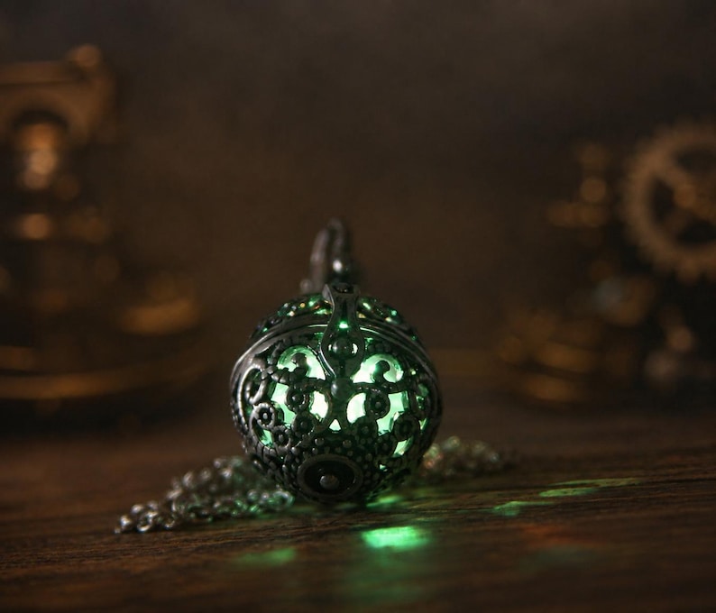 May include: A spherical pendant necklace featuring an intricate, dark metallic filigree cage design. The hollow interior emits a soft, glowing light in a pale green hue, visible through the ornate cutouts of the metal sphere and its chain.
