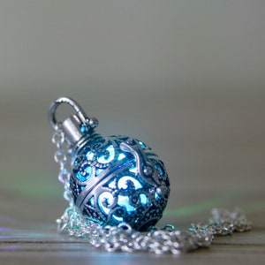 May include: A silver pendant necklace with a blue, ornate, spherical charm. The charm is made of metal with a filigree design and has a glowing blue light inside.