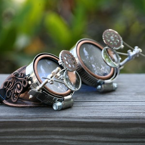 May include: A pair of steampunk goggles with copper and silver accents. The goggles have round lenses and a decorative gear on the side. The goggles are on a wooden surface.