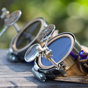 Op de afbeelding: Steampunk-bril met messing en zilveren accenten, met blauw getinte glazen en een vergrootglas aan de zijkant.