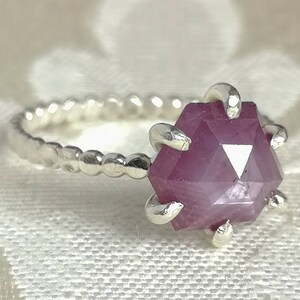 Könnte beinhalten: Ein silberner Ring mit einem rosafarbenen Edelstein, der in einer Krappenfassung sitzt. Der Edelstein ist facettiert und hat eine sechseckige Form.