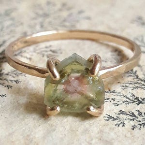 Könnte beinhalten: Ein goldener Ring mit einem grünen und rosafarbenen Edelstein, der in einer dreieckigen Form gefasst ist.