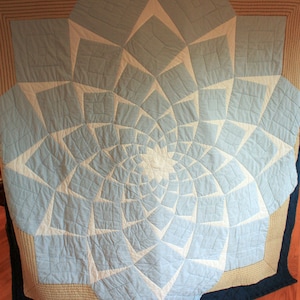 Puede incluir: Una colcha grande, azul y blanca con un patrón en espiral. La colcha está hecha de cuadrados y triángulos, y el patrón en espiral se crea por la forma en que los cuadrados y los triángulos están dispuestos. La colcha está bordeada con un borde marrón y blanco.