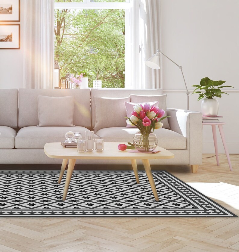May include: A black and white geometric patterned rug with a white border. The rug is in a living room setting with a light gray sofa, a wooden coffee table, and a vase of pink flowers.
