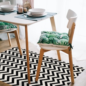 Może przedstawiać: Czarno-biały dywan w chevron pod białym stołem jadalnym z dwoma krzesłami. Jedno krzesło ma zieloną poduszkę z tropikalnym wzorem liści.