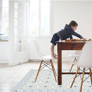 May include: A child playing with toy trains on a dark brown wooden table. The table is surrounded by white chairs with wooden legs. The floor features a patterned rug with blue, gray, and beige floral designs. The room has white walls and a large window.