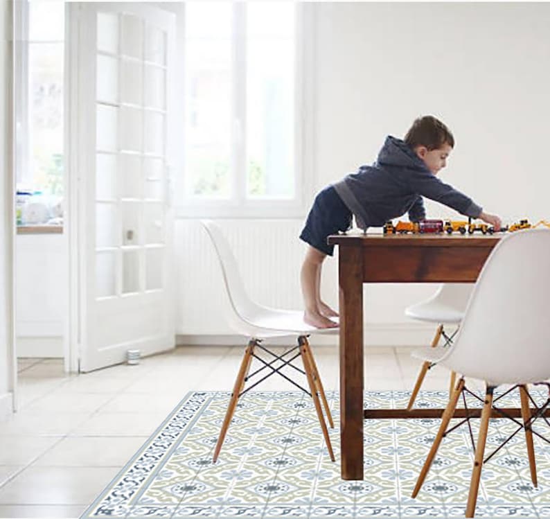 May include: A child playing with toy trains on a dark brown wooden table. The table is surrounded by white chairs with wooden legs. The floor features a patterned rug with blue, gray, and beige floral designs. The room has white walls and a large window.