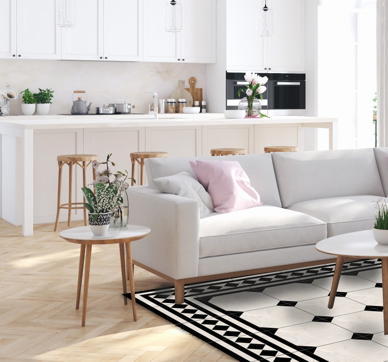 May include: A bright, modern living room with a white sofa, two small round side tables, and a patterned rug. The room features a white kitchen with bar stools and a large window. Decorative plants and flowers add color.
