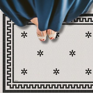 May include: Overhead shot of a patterned tile floor with a person's feet and lower legs. The floor features a white hexagonal tile design with black floral accents and a black border. The person is wearing gold-colored shoes.