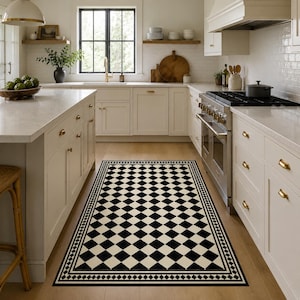 Peut inclure: Un tapis de cuisine rectangulaire à carreaux noirs et crème avec une bordure décorative. Le tapis est posé sur un sol en bois clair dans une cuisine. La cuisine comprend des meubles blancs, une cuisinière et une fenêtre.