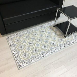 May include: A rectangular patterned rug with a repeating floral design in shades of gray, green, and blue. The rug has a decorative border and is placed on a light-colored wooden floor. The rug is near a black sofa and a small black side table.