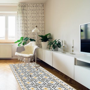 May include: A gray and white patterned area rug with yellow accents. The rug is laid out on a hardwood floor in a living room setting. A white chair with a white sheepskin throw is in the foreground.