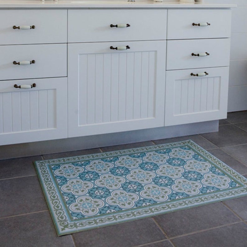 Puede incluir: Una alfombra de ba&ntilde;o rectangular con un patr&oacute;n floral repetido en tonos azules, grises y blancos, enmarcada por un borde verde. La alfombra est&aacute; colocada sobre un suelo de baldosas gris oscuro frente a gabinetes blancos.