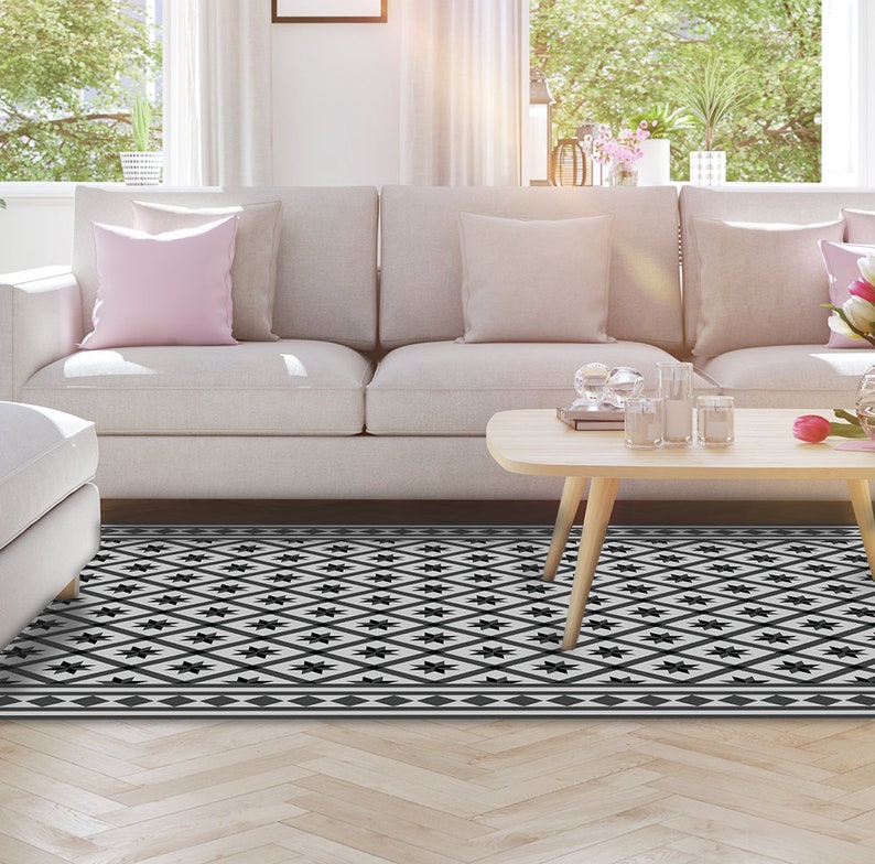 May include: A black and white geometric patterned rug with a diamond design. The rug is placed in front of a white sofa with pink pillows and a wooden coffee table.