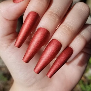 Puede incluir: Un juego de uñas postizas largas, rojas y mate. Las uñas son todas del mismo largo y forma, y tienen un acabado liso y uniforme.