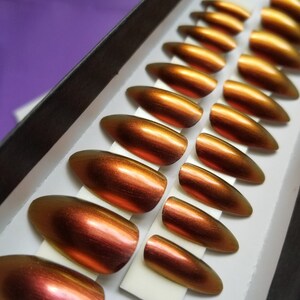 Puede incluir: Un conjunto de 10 uñas postizas en una caja. Las uñas son de color cobre metálico con un efecto degradado. Las uñas están dispuestas en una fila, con la uña más grande a la izquierda y la más pequeña a la derecha.