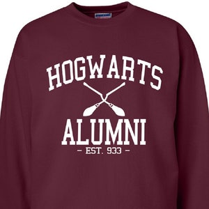 Puede incluir: Sudadera color burdeos con texto blanco que dice "Hogwarts Alumni - Est. 933 -" con un gráfico de dos escobas cruzadas.