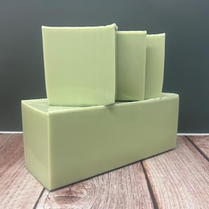 May include: A stack of four light green soap bars. The bottom bar is rectangular, supporting three upright bars. The soap has a smooth, solid appearance. The background is a dark gray, and the surface is a light brown wood.