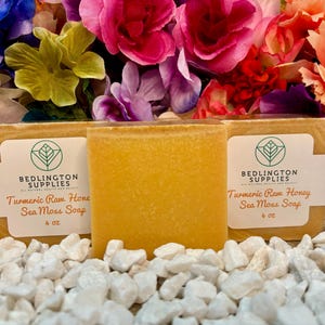 May include: Three bars of yellow turmeric raw honey sea moss soap, each bar is labeled "Bedlington Supplies" and "Turmeric Raw Honey Sea Moss Soap" with a green leaf logo. The soap bars are sitting on a bed of white rocks.