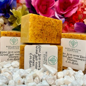 May include: Several bars of yellow soap with dark speckles are stacked on white stones. Each bar is labeled with text that reads "Kojic Acid with Papaya and Turmeric Powder 4 oz Bar". The background features colorful flowers.