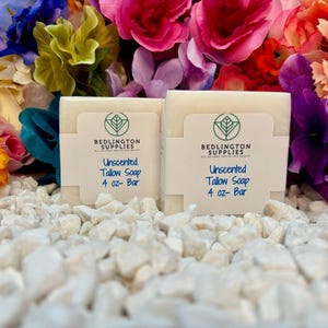 May include: Two white bars of unscented tallow soap, each labeled "Unscented Tallow Soap 4 oz - Bar" and "Bedlington Supplies All Natural Health and Beauty". The soap bars are sitting on a bed of white rocks.