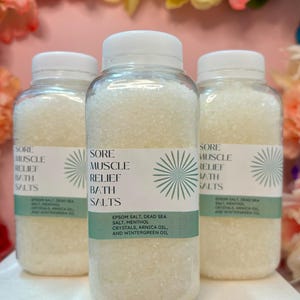 May include: Three clear plastic jars filled with white bath salts. The jars have white lids and green labels with the text "Sore Muscle Relief Bath Salts". The labels also list the ingredients: Epsom salt, Dead Sea salt, menthol crystals, arnica oil, and wintergreen oil.