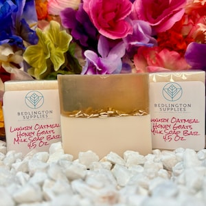 May include: Three bars of handmade soap with a white base and a light brown top. The soap bars are labeled "Luxury Oatmeal Honey Goats Milk Soap Bar 4.5 oz" and are sitting on a bed of white rocks. The soap bars are surrounded by colorful flowers.