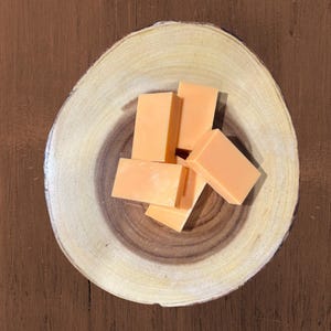 May include: A top-down view of several rectangular, orange-colored soap bars arranged on a round, wooden slice. The wood has a light-colored outer ring and a darker, circular inner pattern. The background is a dark brown surface.