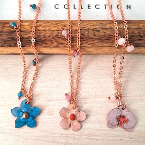 May include: Three necklaces with copper-coloured chains and flower pendants. One is blue, one is pink, and one is lavender. Each necklace has small, colourful beads. The background includes the text "HOME & STYLIN COLLECTION."