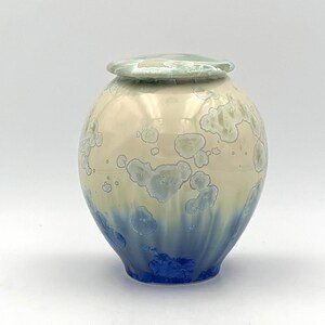 May include: A ceramic urn with a round body and a matching lid. The urn has a colour gradient, shifting from creamy beige at the top to deep blue at the base. The surface is decorated with textured, light blue and white crystalline patterns.