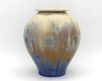 May include: A ceramic urn with a round body and a lid. The urn is decorated with a light brown glaze with blue and white speckles. The bottom of the urn is a deep blue color.