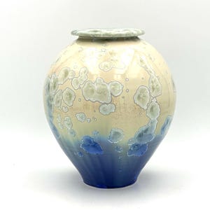 May include: A ceramic vase with a round body and a lid. The vase transitions from a deep blue base to a cream-colored top, with light green and white crystalline patterns. The vase is approximately 25 cm tall.