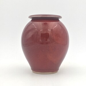 May include: A small, round, red ceramic jar with a lid. The jar has a glossy finish and a slightly textured surface.