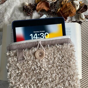 Può includere: Un tablet in una custodia beige e soffice con una parte superiore in velluto a coste e una chiusura con bottone. Lo schermo del tablet mostra l'ora 14:30 e la data, domenica 16 febbraio. La custodia è appoggiata su un libro aperto.