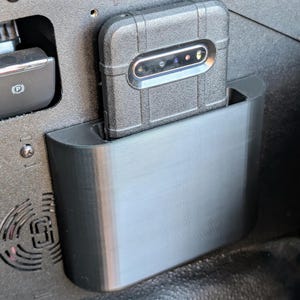 Puede incluir: Una funda de tel&eacute;fono negra con un tel&eacute;fono dentro se encuentra en un soporte de tel&eacute;fono de pl&aacute;stico negro montado en un coche.
